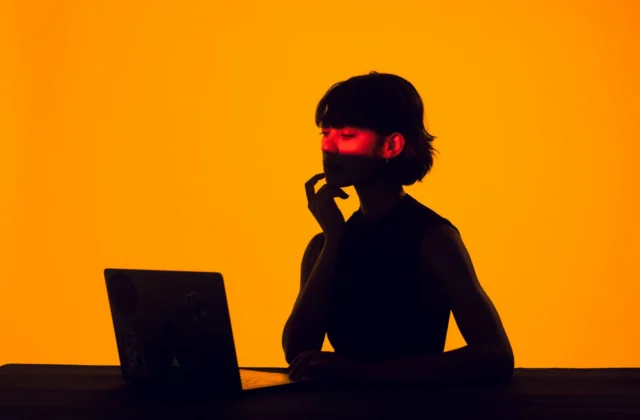 Silhouette of a woman at a laptop against an orange background, a band of red light across her eyes